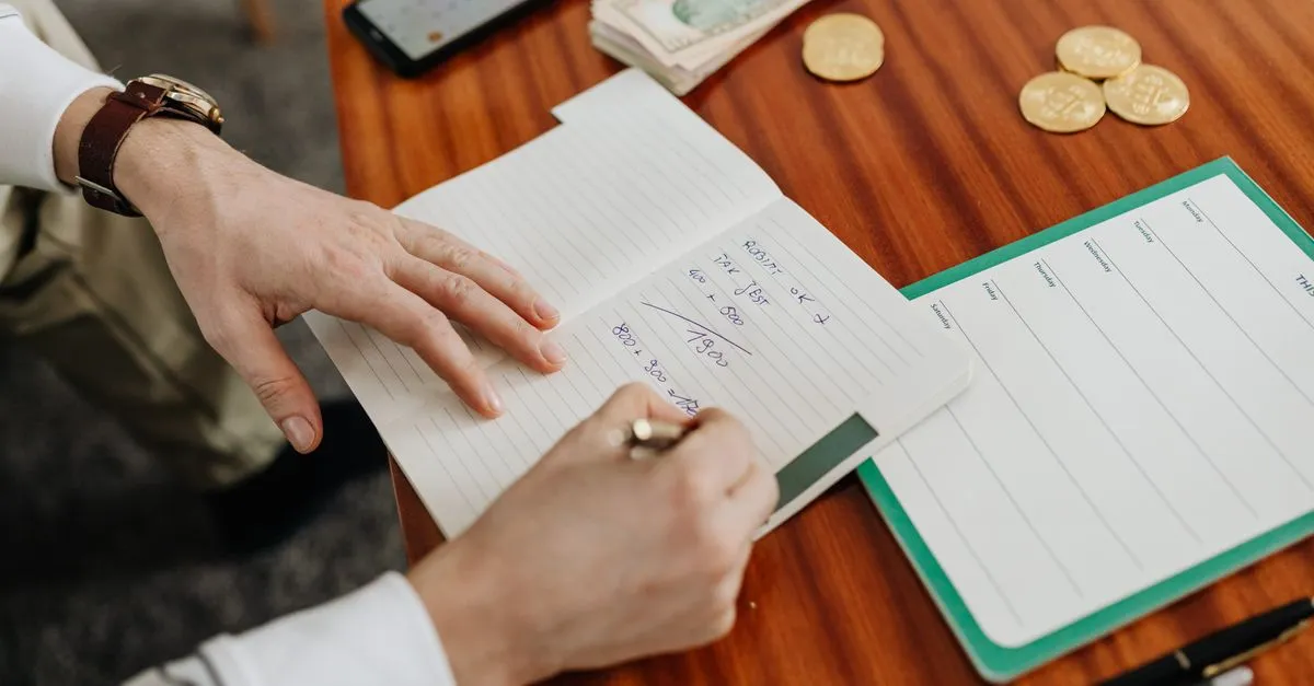 Persona escribiendo su presupuesto a mano en una libreta junto a monedas